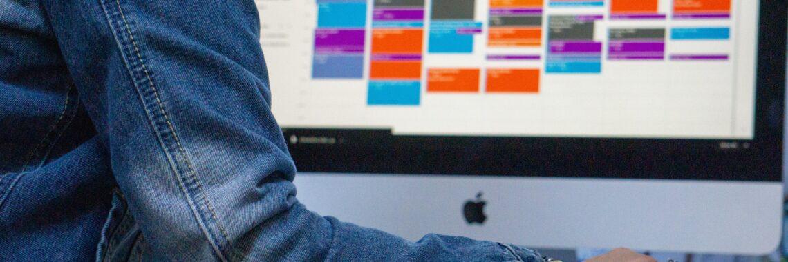 Person in a jean jacket looking at a colorful calendar on a computer monitor.