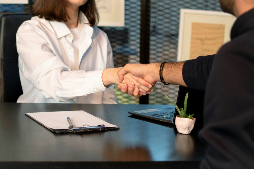 handshake, one woman, one man, over a desk with a laptop and clipboard between them. 